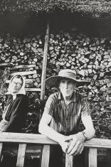 Czarno-biała fotografia portretowa. Przedstawia parę starszych osób na tle składowanego drewna na zewnątrz chaty. Mężczyzna patrzy w stronę obiektywu. Ma pogodną, twarz, nosi słomkowy kapelusz. Kobieta siedzi obok, uśmiecha się, na głowie ma chustę.