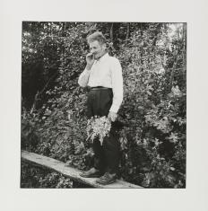Czarno-biała fotografia. Portret starego mężczyzny z papierosem. Postać stoi na drewnianej kładce wśród bujnej roślinności. W ręku trzyma zerwane kwiaty. Widoczna jest  cała sylwetka mężczyzny.