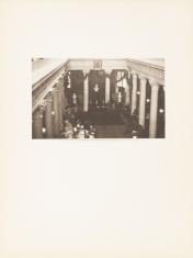 Fotografia auli wykonana w kolorystyce sepii, poziomy kadr. Pomieszczenie sfotografowane z wysokości pierwszego piętra. Wewnątrz widać mężczyzn i kobiety siedzących wokół sali podpartej na białych filarach w stylu korynckim.