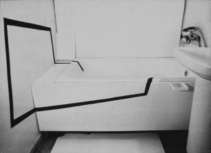 Zdjęcie czarno-białe w układzie poziomym. Na zdjęciu widoczna jest łazienka. Na ścianie i wzdłuż wanny poprowadzona jest czarna linia zaczynająca się w wannie przy węższej krawędzi, później wychodząca na ścianę. Na ścianie linia ta tworzy kształt przypomi