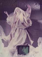 Kobieta w białej, obfitej, zakonnej szacie unosi ręce w górę w geście modlitwy, w fałdach szaty przed nią monitor telewizora, za nią ściana z cegły pomalowana na biało.