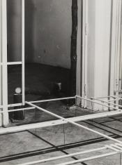 Czarno-białe zdjęcie dokumentacyjne instalacji artystycznej we wnętrzu. Pionowy kadr ujmuje wejście do pokoju zagrodzone pieńkami surowego drewna oraz zawieszoną nad podłogą sieć drewnianych listewek. Konstrukcja rzuca silne cienie na podłogę.