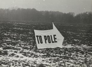 Czarno-biała fotografia w poziomie. Zimowy krajobraz. Pole z partiami przykryte śniegiem. W tle skraj lasu. W centrum podparty transparent w kształcie strzałki z napisem wielkimi literami: To pole.