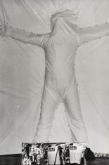 Czarno-białe zdjęcie dokumentacyjne. Pionowy kadr ujmuje pełną sylwetkę człowieka stojącego przy ścianie w rozkroku z uniesionymi w bok ramionami. Postać zakryta jest białym materiałem przypiętym do ściany wokół obrysu ciała. Na dole kolaż 3 innych zdjęć.