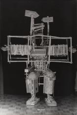 Czarno-białe zdjęcie dokumentacyjne, pionowy kadr. Drewniana konstrukcja, jakby kostium. Stelaż zaprojektowany w pozycji siedzącej z uniesionymi w bok ramionami. Zamiast postaci umieszczone są odlewy stóp i dłoni. 