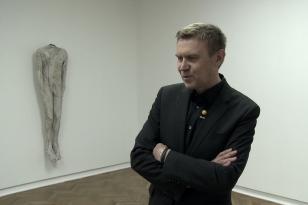 W sali wystawowej stoi ubrany na czarno artysta Mirosław Bałka ze skrzyżowanymi przed sobą rękami, za nim na ścianie wisi rzeźba - naturalnej wielkości bezgłowe ciało mężczyzny.