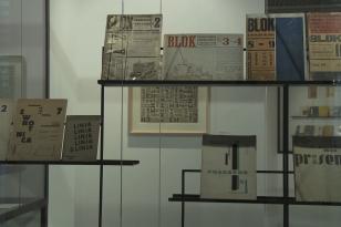Na metalowej ażurowej półce ustawione są różnorodne awangardowe druki, w tle na ścianie oprawiona praca liternicza.