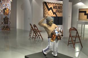 W sali wystawienniczej na środku stoi na postumencie rzeźba - postać niskiego mężczyzny z uniesioną ręką zaciśniętą w pięść i naręczem ubrań w drugiej, ma czapkę i okulary, w tle inne prace i krzesła.