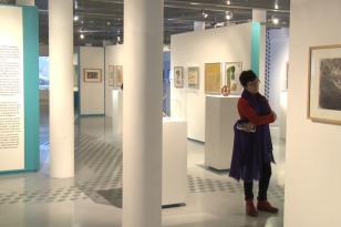 Biała sala wystawowa z niebieskimi akcentami, wypełniona kolumnami i kubikami do prezentacji prac, na ścianach obrazy, po prawej stoi kobieta oglądająca wystawę.