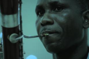 W dużym zbliżeniu twarz czarnoskórego mężczyzny grajacego na instrumencie dętym, ma wydęte policzki, całość pogrążona w niebieskim śwetle. 