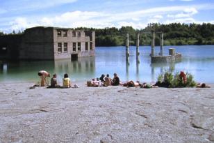 Na brzegu zbiornika wodnego siedzi grupa plażujących osób, w wodzie przed nimi zatopiony częściowo dom i fragmenty betonowych konstrukcji. 