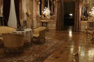 Widok na bogato zdobione wnętrze salonu, po lewej stolik i trzy fotele, na ścianach flankujących wejście lustra z bogatą dekoracją, w narożniku wazon na postumencie.