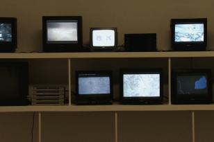 Na dwóch przyścienych półkach ostawione są liczne telewizory, ich jasno rozświetlone ekrany kontrastują z ciepłym, beżowym tłem.