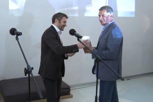 Na scene dwóch mężczyzn i dwa mikrofony, są zwróceni twarzami do siebie, jeden przekazuje drugiemu nagrodę w postaci białego kubika.  