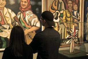 Dwie postacie widzów wystawy stojące tyłem, przed nimi oglądane prace - trzy duże, kolorowe obrazy figuratywne, po prawej w  gablocie rzeźba.