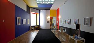 Kolorowe zdjęcie przedstawiające wnętrze Sali Neoplastycznej - sufit i ściany boczne w kolorach niebieskim, czerwonym, żółtym i niekolorach szarym, czarnym i białym, wiszą obrazy abstrakcyjne, w głębi czarno-żółte proste meble. 