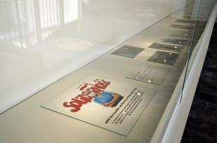 Ekspozycja materiałów w szklanej gablocie: płaskie, kolorowe wydruki prac uczestników projektu Praca = Dokumentacja Wyobraźni, na pierwszym planie fotografia Sanji Ivekowić „Solidarność 1989-2020” 