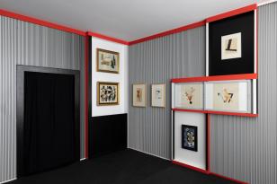 Kolorowe zdjęcie sali wystawienniczej zorientowane poziomo; widok na róg pomieszczenia z drzwiami po lewo;  na ścianie prawej i  w rogu po lewej stronie wisi 8 abstrakcyjnych prac w czerwonych ramach;  tapeta jest w czarno-białe pionowe pasy.
