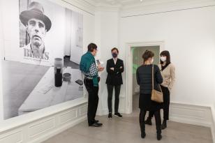 Widok ogólny. Mniejsza sala galerii. Na ścianie czarno-biała fototapeta z wizerunkiem Josepha Beuysa w kapeluszu oraz goście wernisażu (4 osoby). 