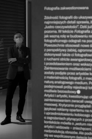 Kierownik Działu Komunikacji Adam Jurek. Postać w garniturze, stojąca w głębi pomieszczenia, na pierwszym planie niewyraźny tekst objaśniający wystawę umieszczony na ścianie