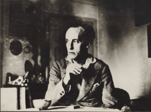 Władysław Strzemiński siedzi przy stole, w ręce trzyma papierosa, w tle mieszkanie. 