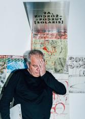 Autor wystawy Marek Sobczyk na tle swojego obrazu o kształcie rozłożonego sześcianu, każda część opowiada inną historię. Artysta uchwycony w ekspresyjnym geście, przykłada lewą dłoń do twarzy 