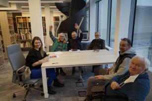 Członkowie grupy Kódź Kaliska siedzą przy prostokątnym stole w czytelni biblioteki, z lawej na fotelu Małgorzata Chmiel. Adam Rzepecki i Andrzej Świetlik podnoszą do góry ręce, pozdrawiając kogoś, kto wchodzi do biblioteki