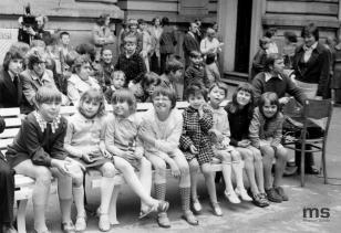 Fotografia czarno-biała przedstawiająca grupę dzieci na dziedzińcu muzeum, siedzących na ławkach i stojących za nimi, uśmiechajacych się. 