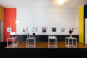 Widok sali wystawowej - na białej ścianie z czerwonym pasem po lewej stronie rzędem wisi 7 obrazów w formacie zbliżonym do kwadratu,  na pierwszym planie 4 rzeźby na postumentach, z prawej kawałek ściany  w kolorze żółtym.