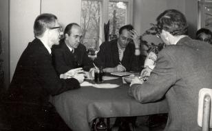 Na czarnobiałej fotografii przy stole siedzi 5 osób, na pierwszym planie mężczyzna tyłem, mężczyzna z lewej profilem, dwie osoby en face, jedna widoczna tylko zza ramienia osoby na pierwszym planie. 