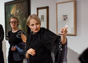 Kuratorka wystawy Dorota Monkiewicz, w głębi Karolina Dróżdż (Dział Komunikacji). Obie panie stoją na tle trzech obrazów zawieszonych na ścianie. Kuratorka tłumacząc, wskazuje na coś ręką