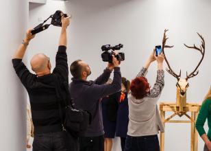 Scena z wernisażu. Troje ludzi trzyma ręce wysoko uniesione do góry, trzymając w nich aparaty, kamerę. Z prawej strony rzeźba składający się z głowy jelenia z ogromnym porożem. Głowa przytwierdzona jest do drewnianego korpusu