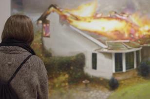 Na zdjęciu kobieta w płaszczu i z plecakiem odwrócona tyłem do nas, a przodem do dużego obrazu, przedstawiającego dom stojący w płomieniach. 