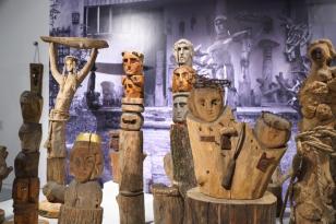 Grupa drewnianych rzeźb przypominających tradycyjne świątki ustawiona na tle wielkoformatowej fotografii przedstawiającej rzeźby w otoczeniu domu artysty.
