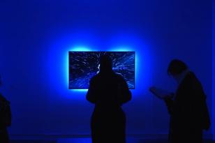 Cały kadr zatopiony w niebieskim świetle, przed ekranem wiszącego na ścianie monitora stoją dwie osoby, oglądając pracę.