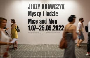 Zdjęcie w hallu budynku ms2. Na ścianie wielkimi literami wypisany tytuł wystawy w języku polskim i angielskim oraz daty jej trwania. Przez kadr przewijają się postaci widzów przybyłych na wernisaż