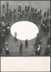 Kadr przedstawia duży biały okrąg, który położony został na trotuarze ułożonym z graniastych płyt. Gromadzą się wokół niego ludzie, niektórzy  trzymają w rękach parasole