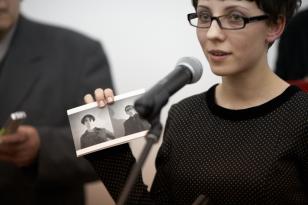 Zbliżenie na młodą kobietę w okularach, z krótkimi włosami, która stoi przy mikrofonie i demonstruje widzom zaproszenie na wystawę - portret podwójny dyrektora Stanisławskiego i Urszuli Czartoryskiej