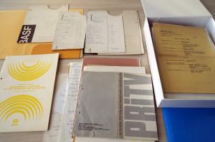Na blacie stołu leżą porozkładane archiwalia: teczki, karty maszynopisów, wydawnictwa, część z nich leży w tekturowym, prostokątnym pudle z otwieraną przykrywką
