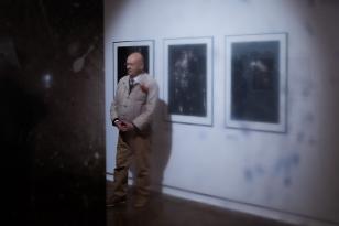 Mężczyzna z ogoloną głową stoi na tle trzech wiszących pionowo fotografii. Obraz wykonany jest przez folię, która częściowo zasłania lub rozmywa detale