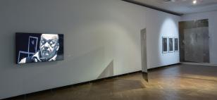 W dużej sali wiszą wielkie prostokątne płaszczyzny zadrukowane abstrakcyjnymi kompozycjami. Prace są przezroczyste, wykonane z plastiku.  Widać kilka prac oświetlonych okrągłą poświatą i telewizor z kadrem - twarzą mężczyzny