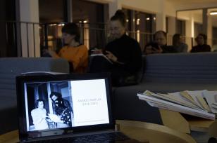 Na niskim stoliku stoi otwarty laptop. Na ekranie zaproszenie na wystawę: zdjęcie Andrzeja Partuma w towarzystwie Ewy Partum, obok imię i nazwisko artysty i daty biograficzne. W niewyraźnym tle wnętrze pomieszczenia i sylwetki widzów