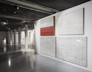 Z prawej strony kadru na dobudowanej ścianie zawieszone są cztery prostokątne, duże obrazy abstrakcyjne. Białe. Pokryte są gęsto znakami ułożonymi w linie. Jeden z obrazów wzbogacony jest o kolor czerwony. Obraz układa się w polską flagę