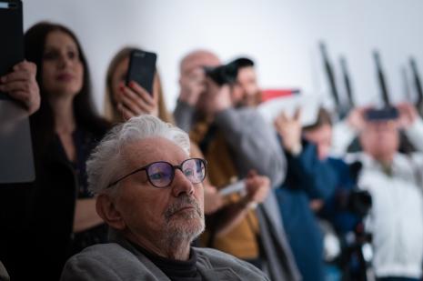 Prof. Jerzy Treliński (b. wykładowca ASP w Łodzi)
Na pierwszym planie znajduje się starszy mężczyzna z siwymi włosami, bródką, w okularach. Słucha, siedząc na niewidocznym wózku inwalidzkim. W głębi nieostre sylwetki uczestników wernisażu