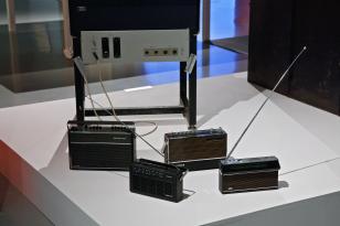 Na niskim kwadratowym podeście stoją cztery przenośne radioodbiorniki z lat 80-tych XX wieku. Są stosunkowo niewielkie, mają głośnik, pokrętła i szklaną skalę radiową oraz wysuwane długie anteny