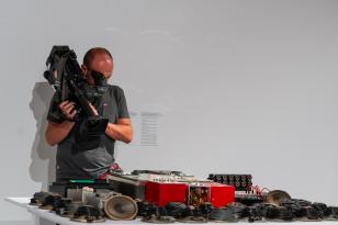 Kamerzysta z dużą kamerą na ramieniu filmuje z bliska pracę umieszczoną na stole. Składa się ona z szeregu części głośników, wymontowanych membran i przewodów, magnetofonu, przetworników, konsoli