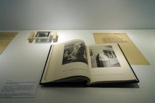 Zbliżenie na eksponaty znajdujące się w punktowo oświetlonej gablocie. Na pierwszym planie leży otwarta książka z czarno-białymi ilustracjami, wyżej znajduje się pożółkły dokument i na szklanej szybce wspartej na drewnianych klockach szklany negatyw