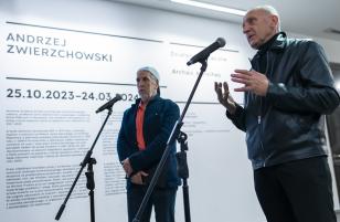 Zdjęcie wykonane lekko z dołu. Dwóch mężczyzn w sile wieku stoi przy statywach z mikrofonami. Mężczyzna z prawej, posiwiały, w czarnej skórzanej kurtce przemawia. Gestykuluje. Drugi z mężczyzn obserwuje publiczność