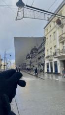 Układ pionowy. Z lewego dolnego narożnika wysatje dłon trzymająca czarno-białą fotografię. W tle za fotografią widać ulicę i otaczające kamienice.