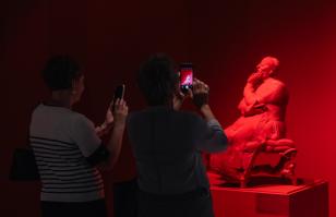 Układ poziomy. Po prawej stronie w tle czerwona rzeźba na czerwonym postumencie. Przed rzeźbą po lewej stronie sylwetki 2 kobiet stojących tyłem do kadru, Fotografują dzieło..